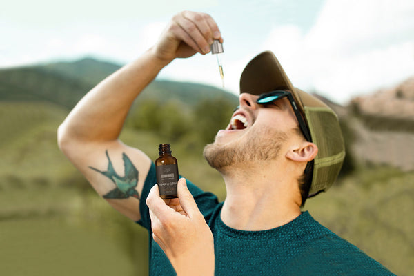 Man with hat and tattoo standing in field taking Amphora CBD oil drops uk.