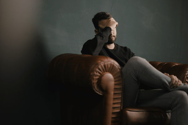 A man sitting in a sofa, cross-legged, palming his face. CBD to aid mood  